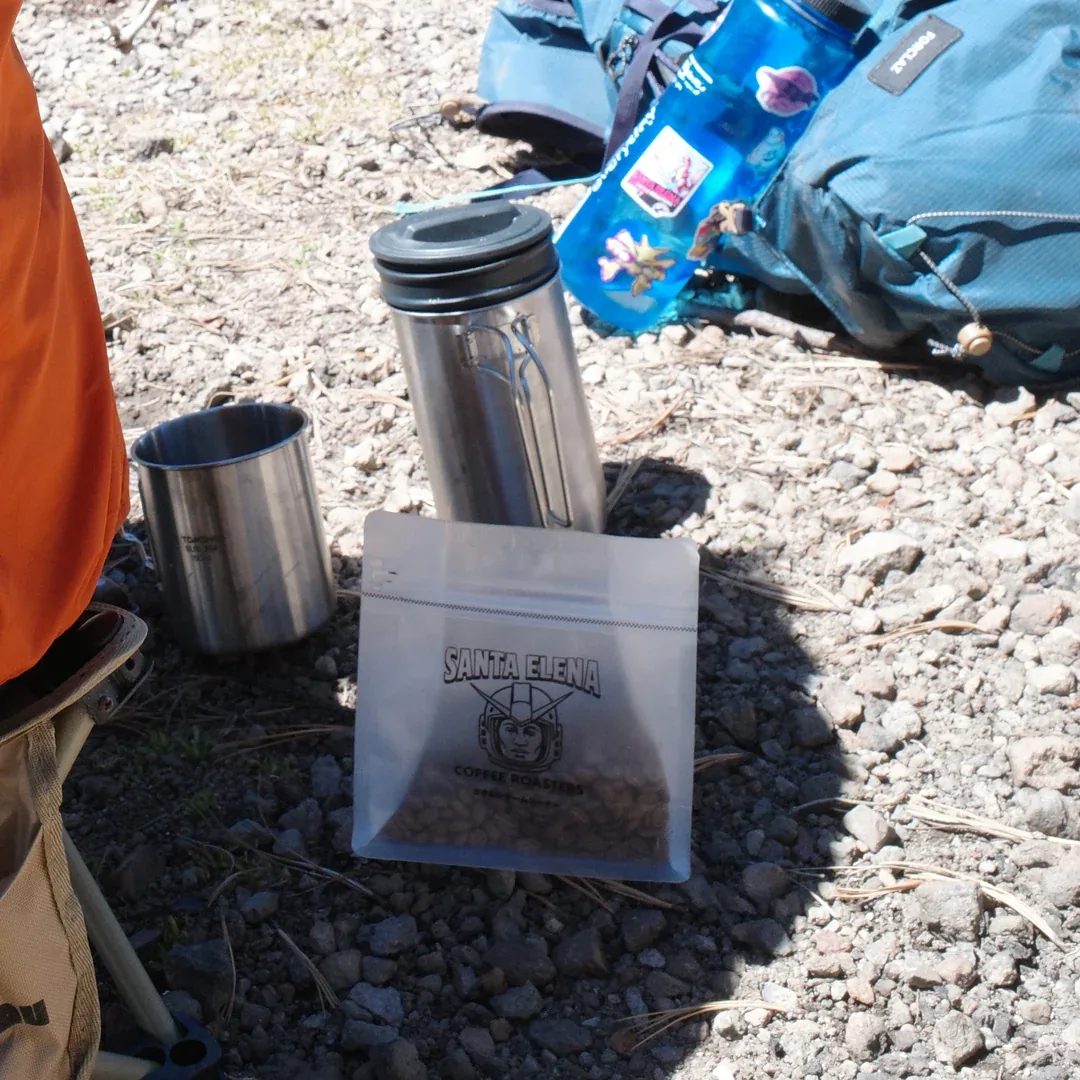 Bolsa de café Geisha Santa Elena con granos junto a una prensa y taza de acero en un día de campo