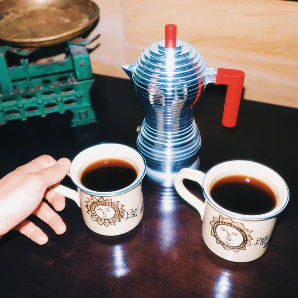 Cafetera Alessi Pulcina roja junto a dos tazas de café servido sobre una mesa de madera