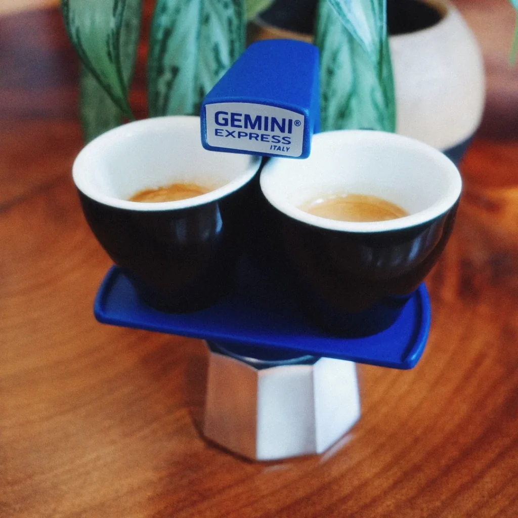 Cafetera Gemini Express azul sirviendo dos espressos en tazas negras, con una planta al fondo