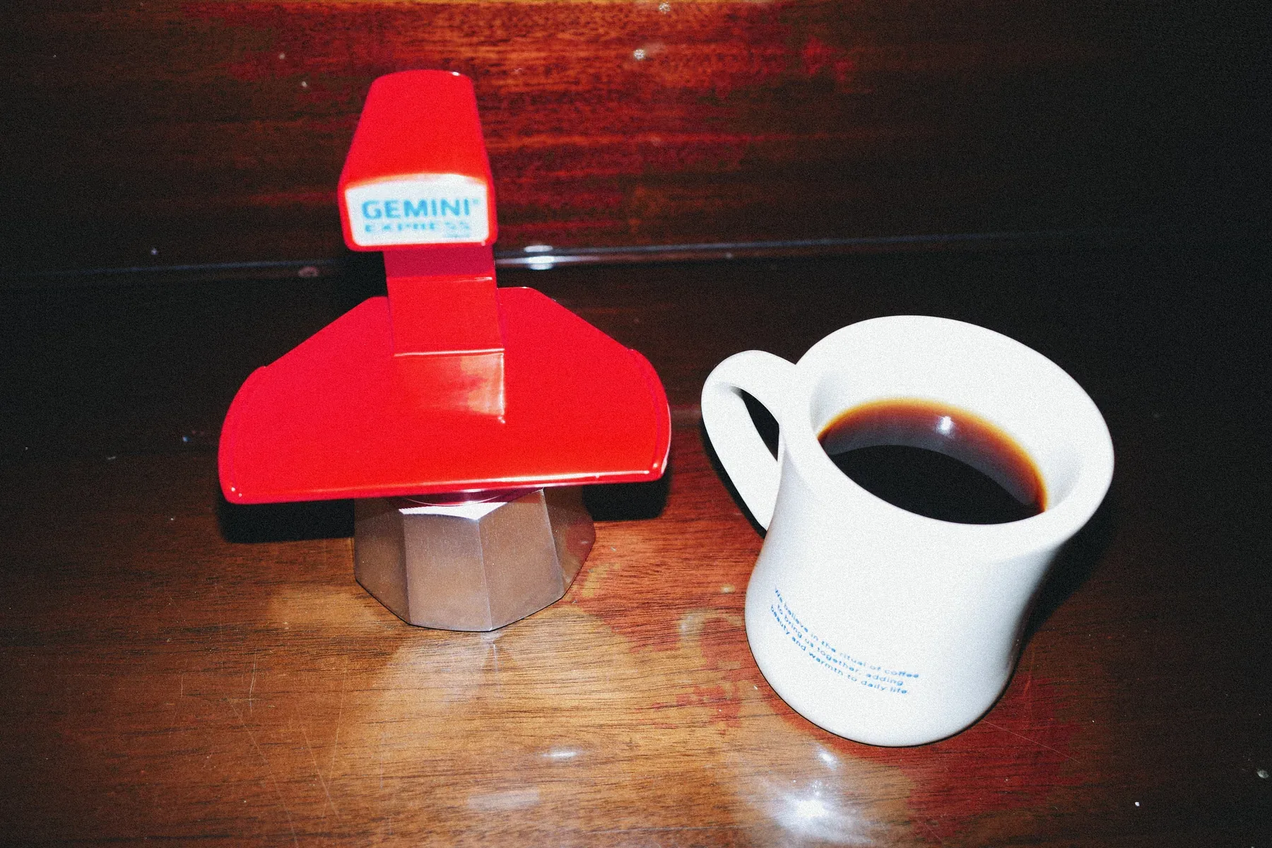 Cafetera Gemini Express roja y una taza de café sobre una mesa de madera